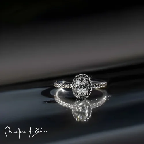 Anillo de compromiso ovalado con piedras, reflejándose en una superficie brillante.