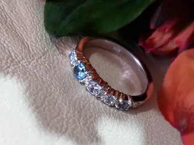 Anillo plateado #2213 con topacio azul central y línea de cristales brillantes sobre fondo claro con elementos florales rojos