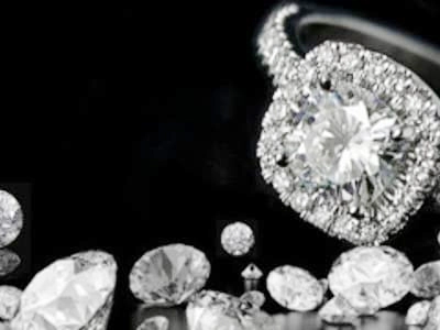 Imagen en blanco y negro sobre fondo negro. En primer plano, a la derecha, se muestra un anillo de compromiso con un diamante central rodeado por un halo de diamantes más pequeños en un engaste cuadrado. En la parte inferior de la imagen, se observan varios diamantes sueltos de diferentes tamaños y cortes, reflejando la luz.