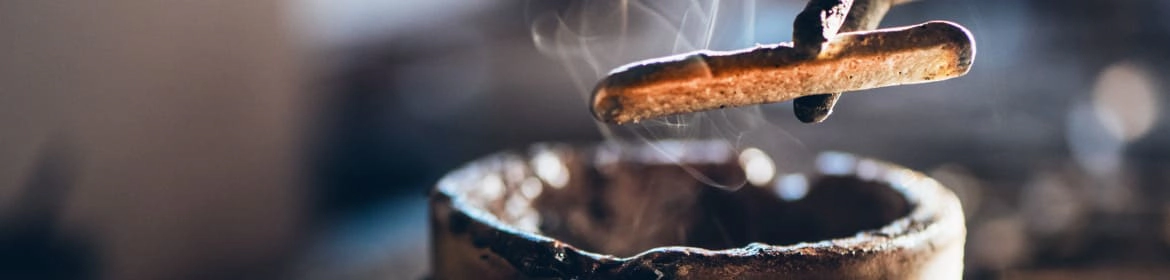 Imagen que muestra un molde de fundición con metal caliente, con vapor saliendo, mientras se utiliza una herramienta para extraer el metal fundido en un entorno de trabajo artesanal.
