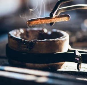 Imagen que muestra un molde de fundición con metal caliente, con vapor saliendo, mientras se utiliza una herramienta para extraer el metal fundido en un entorno de trabajo artesanal.