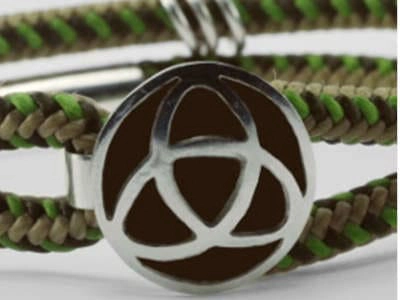 Primer plano de una pulsera hecha a mano con dos hebras de cordón trenzado en tonos tierra y verde. Las hebras están unidas por un broche rectangular de metal plateado y un adorno circular que presenta un diseño de triquetra recortado, revelando un fondo oscuro contrastante. El enfoque nítido en la pulsera resalta la artesanía y los materiales utilizados.