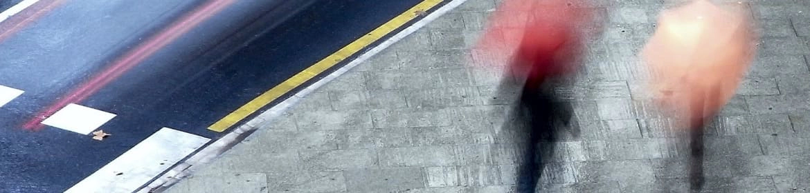 Una vista aérea de dos personas que caminan por una acera con paraguas en un día lluvioso. La acera está hecha de adoquines grises y hay una calle con un coche que pasa rápidamente en la parte superior de la imagen. Los paraguas son de color rojo y melocotón, y las personas que los sostienen están borrosas por el movimiento, lo que sugiere que caminan a un ritmo rápido. La imagen transmite una sensación de movimiento y la atmósfera de un día lluvioso en un entorno urbano.
