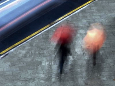 Una vista aérea de dos personas que caminan por una acera con paraguas en un día lluvioso. La acera está hecha de adoquines grises y hay una calle con un coche que pasa rápidamente en la parte superior de la imagen. Los paraguas son de color rojo y melocotón, y las personas que los sostienen están borrosas por el movimiento, lo que sugiere que caminan a un ritmo rápido. La imagen transmite una sensación de movimiento y la atmósfera de un día lluvioso en un entorno urbano.