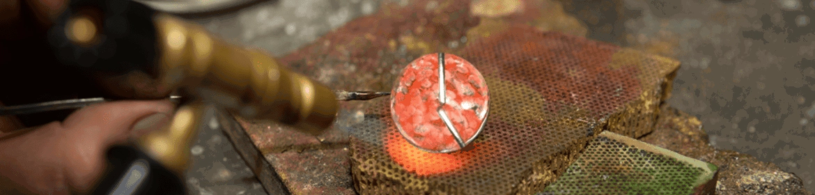 Un joyero utiliza un soplete para soldar un bisel de plata alrededor de una cabujón de ópalo rosa. El ópalo se coloca sobre una superficie resistente al calor y el joyero sujeta el bisel en su lugar con unas pinzas. La llama del soplete es pequeña y precisa, y el joyero la mueve cuidadosamente alrededor del bisel para calentar la plata hasta el punto de fusión. La plata fundida fluye alrededor del ópalo, creando un ajuste seguro y permanente.