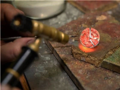 Un joyero utiliza un soplete para soldar un bisel de plata alrededor de una cabujón de ópalo rosa. El ópalo se coloca sobre una superficie resistente al calor y el joyero sujeta el bisel en su lugar con unas pinzas. La llama del soplete es pequeña y precisa, y el joyero la mueve cuidadosamente alrededor del bisel para calentar la plata hasta el punto de fusión. La plata fundida fluye alrededor del ópalo, creando un ajuste seguro y permanente.
