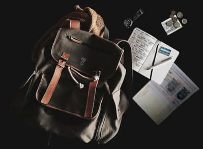  Imagen que muestra una mochila de cuero con detalles en cuero marrón, acompañada de una libreta, un bolígrafo, monedas y documentos de viaje, sobre un fondo oscuro, creando una atmósfera de aventura y exploración.