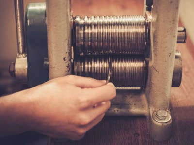 Mano ajustando una lámina de metal en una máquina de laminado, mostrando el mecanismo de compresión de rodillos metálicos en un entorno de trabajo artesanal.