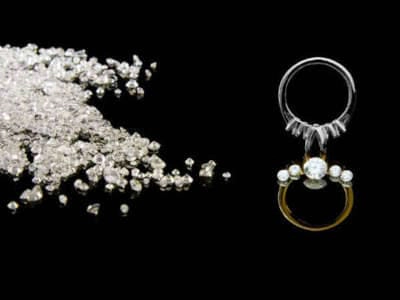 Imagen sobre fondo negro mostrando a la izquierda un montón de diamantes en bruto dispersos, y a la derecha un anillo de joyería elaborado. El anillo presenta un diseño de dos aros: uno superior plateado con pequeños diamantes engastados, y uno inferior dorado con un diamante central más grande rodeado de perlas o diamantes más pequeños