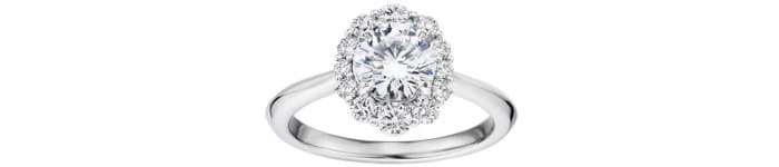 Este anillo de compromiso presenta una montura de halo que rodea un diamante central de corte redondo brillante. La montura es de metal preciosa, probablemente oro blanco, con un acabado pulido y brillante. El diseño de halo cuenta con pequeños diamantes rodeando el diamante central, lo que crea un efecto visual de un diamante de mayor tamaño. Este tipo de montura realza y enmarca el diamante central, convirtiéndolo en el elemento focal del anillo. Es un diseño clásico y elegante que ofrece un aspecto suntuoso y refinado.