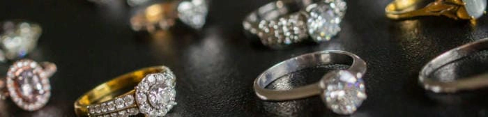 Varios anillos de diamantes de diferentes estilos y metales preciosos se muestran sobre un fondo negro texturizado. Los anillos presentan una variedad de engastes, incluidos engastes de halo, engastes de solitario y engastes de pavé, mostrando diferentes formas y tamaños de diamantes. La imagen está enfocada selectivamente en algunos de los anillos, creando una sensación de profundidad y resaltando el brillo de los diamantes.