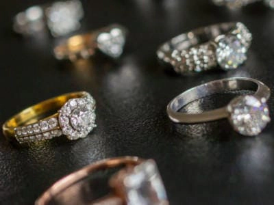 Varios anillos de diamantes de diferentes estilos y metales preciosos se muestran sobre un fondo negro texturizado. Los anillos presentan una variedad de engastes, incluidos engastes de halo, engastes de solitario y engastes de pavé, mostrando diferentes formas y tamaños de diamantes. La imagen está enfocada selectivamente en algunos de los anillos, creando una sensación de profundidad y resaltando el brillo de los diamantes.