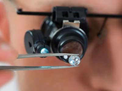 Un gemólogo utiliza una lupa con luz incorporada para examinar un diamante en bruto sostenido con unas pinzas. El rostro del gemólogo está desenfocado, lo que centra la atención en el diamante y las herramientas utilizadas para su inspección. La lupa permite una visión ampliada de la piedra, facilitando la evaluación de su claridad, color y otras características importantes.