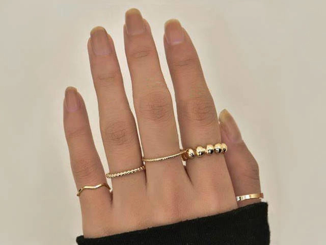 Mano mostrando varios anillos de oro en diferentes estilos, incluyendo diseños minimalistas y detalles decorativos, sobre un fondo neutro.