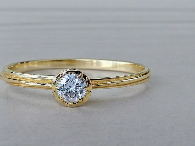 Un anillo de oro con un diamante solitario engastado en bisel se muestra sobre un fondo liso y de color claro. El anillo tiene una banda de oro simple con un diseño de doble línea que envuelve el dedo. El diamante es redondo y brillante, y está asegurado por un engaste de bisel circular que lo rodea por completo. La imagen está enfocada en el anillo, destacando el brillo del diamante y el pulido del oro.