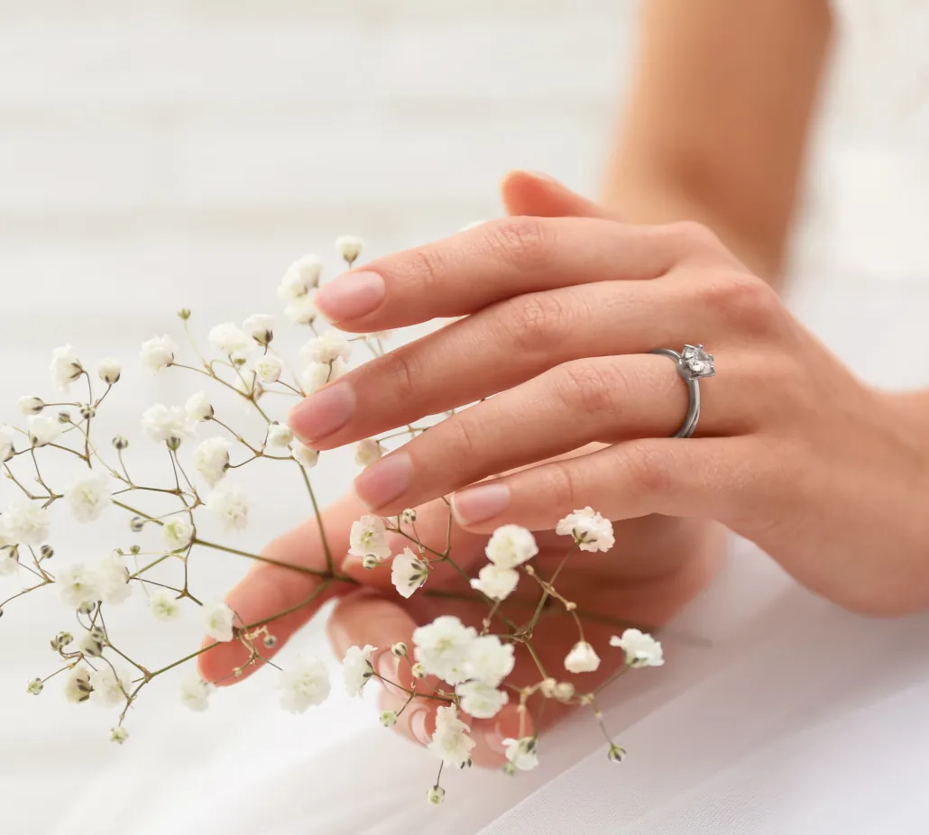 Primer plano de una mano femenina con un anillo de compromiso solitario de diamante, sosteniendo delicadas flores blancas de gypsophila (o paniculata). Las uñas están arregladas con un esmalte nude natural, y la imagen transmite un ambiente romántico y etéreo sobre un fondo claro.